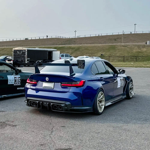 Blue BMW G80 M3 with aggressive carbon fiber aero kit, front splitter, vented fenders, and side skirts parked beside a professional race track during sunset.