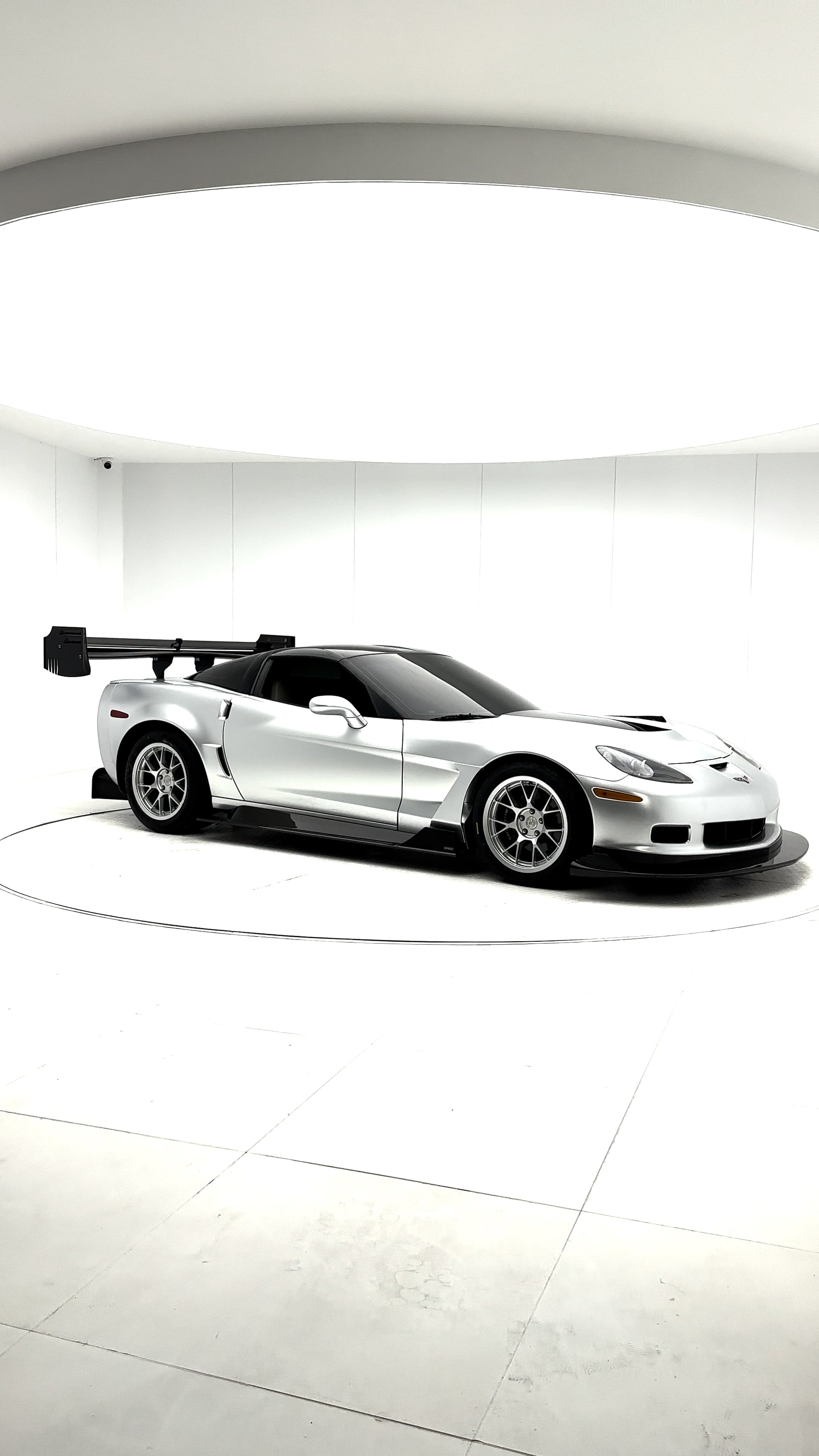 Close-up of a silver wide-body Chevrolet Corvette performance car displayed indoors, featuring aggressive aerodynamic bodywork, carbon fiber front splitter, vented hood, and lightweight multi-spoke wheels. The image highlights the custom motorsport styling and premium carbon fiber aero components under exhibition lighting.