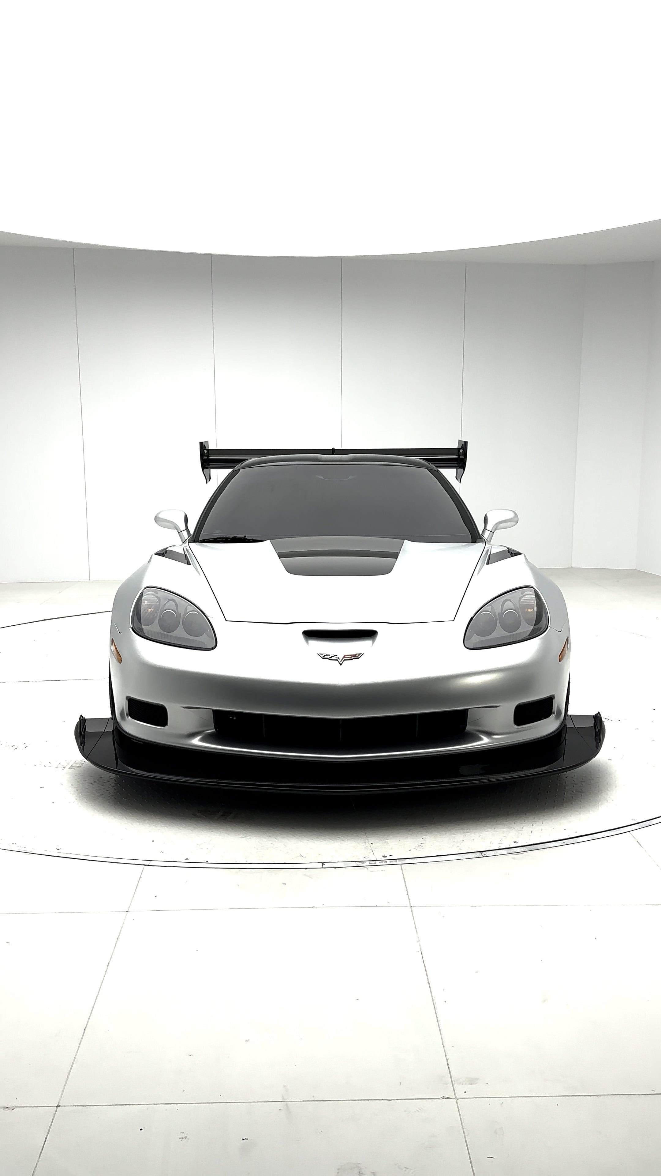 Close-up of a silver wide-body Chevrolet Corvette performance car displayed indoors, featuring aggressive aerodynamic bodywork, carbon fiber front splitter, vented hood, and lightweight multi-spoke wheels. The image highlights the custom motorsport styling and premium carbon fiber aero components under exhibition lighting.