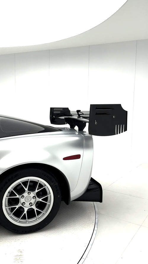 Close-up of a silver wide-body Chevrolet Corvette performance car displayed indoors, featuring aggressive aerodynamic bodywork, carbon fiber front splitter, vented hood, and lightweight multi-spoke wheels. The image highlights the custom motorsport styling and premium carbon fiber aero components under exhibition lighting.