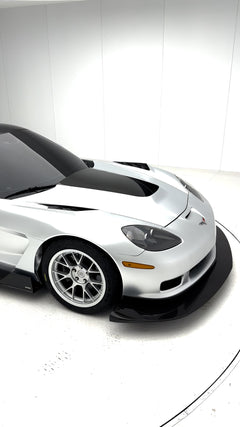 Close-up of a silver wide-body Chevrolet Corvette performance car displayed indoors, featuring aggressive aerodynamic bodywork, carbon fiber front splitter, vented hood, and lightweight multi-spoke wheels. The image highlights the custom motorsport styling and premium carbon fiber aero components under exhibition lighting.
