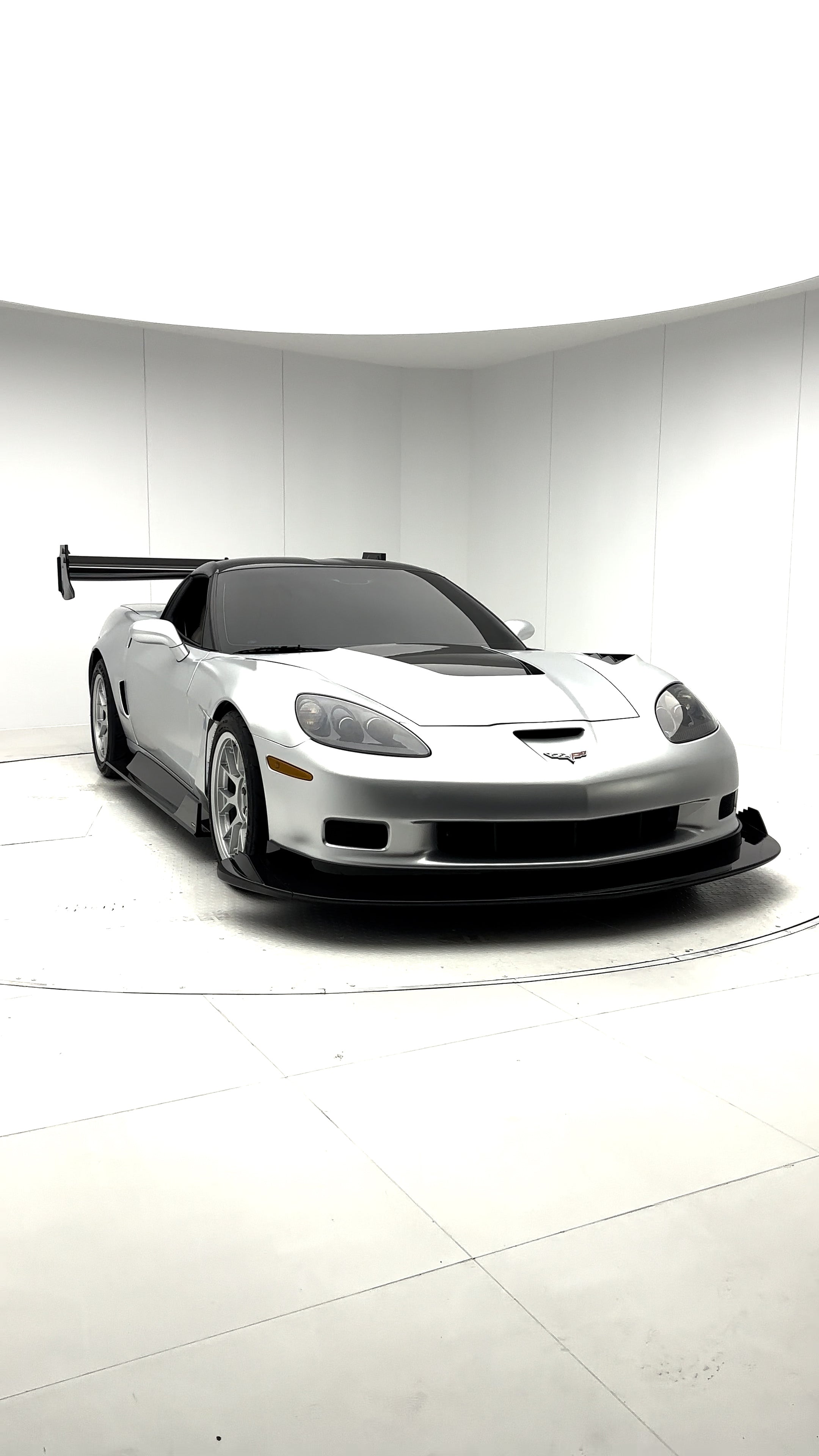 Close-up of a silver wide-body Chevrolet Corvette performance car displayed indoors, featuring aggressive aerodynamic bodywork, carbon fiber front splitter, vented hood, and lightweight multi-spoke wheels. The image highlights the custom motorsport styling and premium carbon fiber aero components under exhibition lighting.