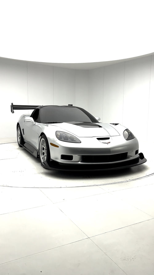Close-up of a silver wide-body Chevrolet Corvette performance car displayed indoors, featuring aggressive aerodynamic bodywork, carbon fiber front splitter, vented hood, and lightweight multi-spoke wheels. The image highlights the custom motorsport styling and premium carbon fiber aero components under exhibition lighting.