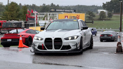 White BMW G80 M3 race car with REVOZPORT carbon fiber aero kit, front splitter, canards, and large rear wing on a wet track paddock during a motorsport event.