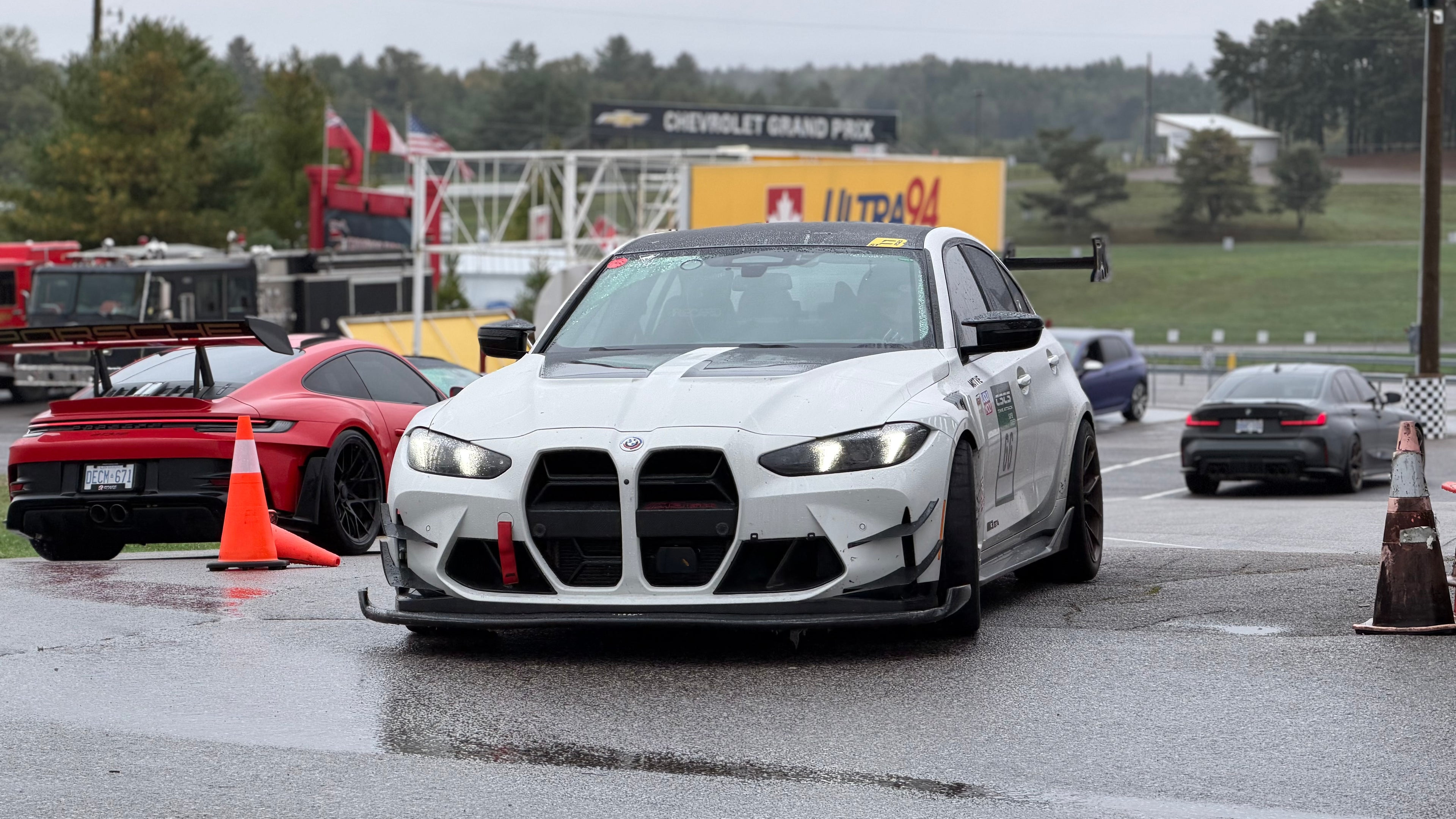 White BMW G80 M3 race car with REVOZPORT carbon fiber aero kit, front splitter, canards, and large rear wing on a wet track paddock during a motorsport event.