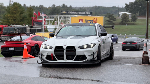 White BMW G80 M3 race car with REVOZPORT carbon fiber aero kit, front splitter, canards, and large rear wing on a wet track paddock during a motorsport event.