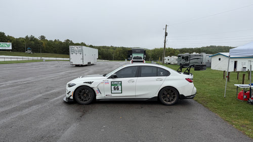 White BMW G80 M3 race car with REVOZPORT carbon fiber aero kit, front splitter, canards, and large rear wing on a wet track paddock during a motorsport event.