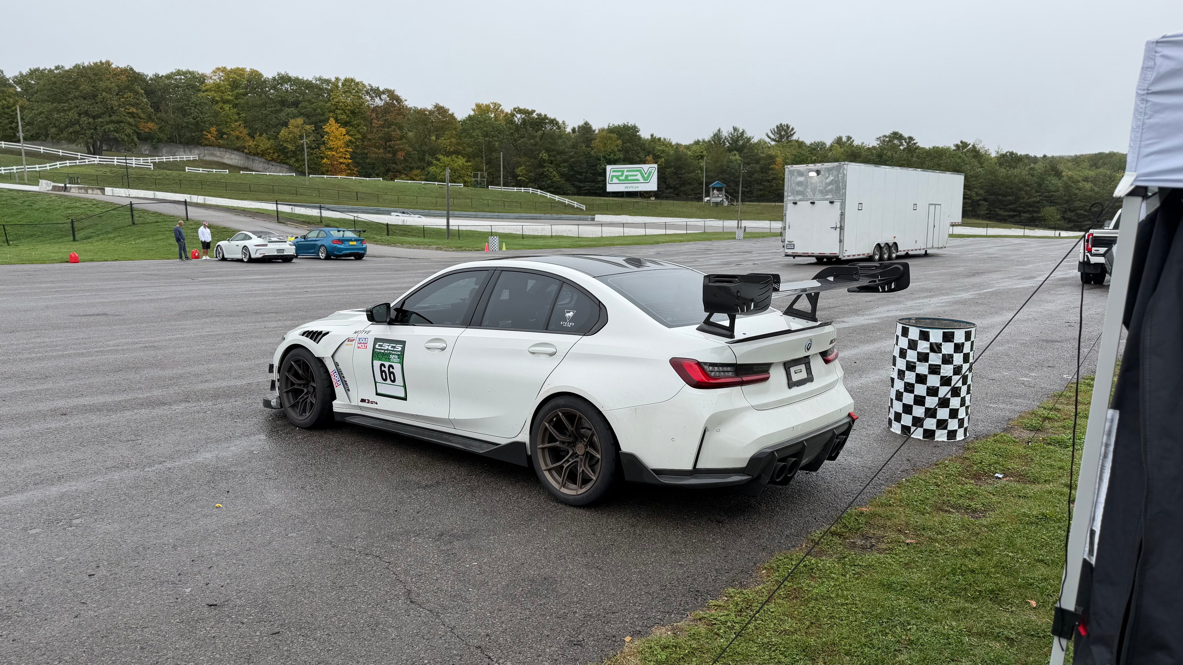 White BMW G80 M3 race car with REVOZPORT carbon fiber aero kit, front splitter, canards, and large rear wing on a wet track paddock during a motorsport event.