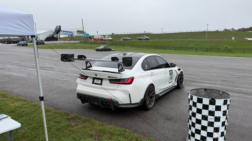 White BMW G80 M3 race car with REVOZPORT carbon fiber aero kit, front splitter, canards, and large rear wing on a wet track paddock during a motorsport event.