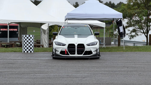 White BMW G80 M3 race car with REVOZPORT carbon fiber aero kit, front splitter, canards, and large rear wing on a wet track paddock during a motorsport event.