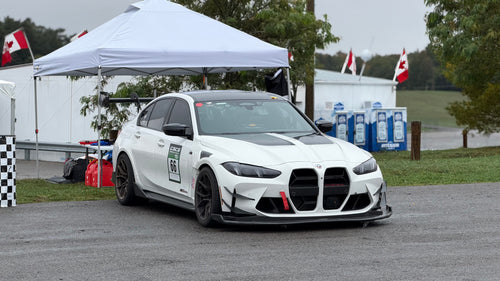 White BMW G80 M3 race car with REVOZPORT carbon fiber aero kit, front splitter, canards, and large rear wing on a wet track paddock during a motorsport event.