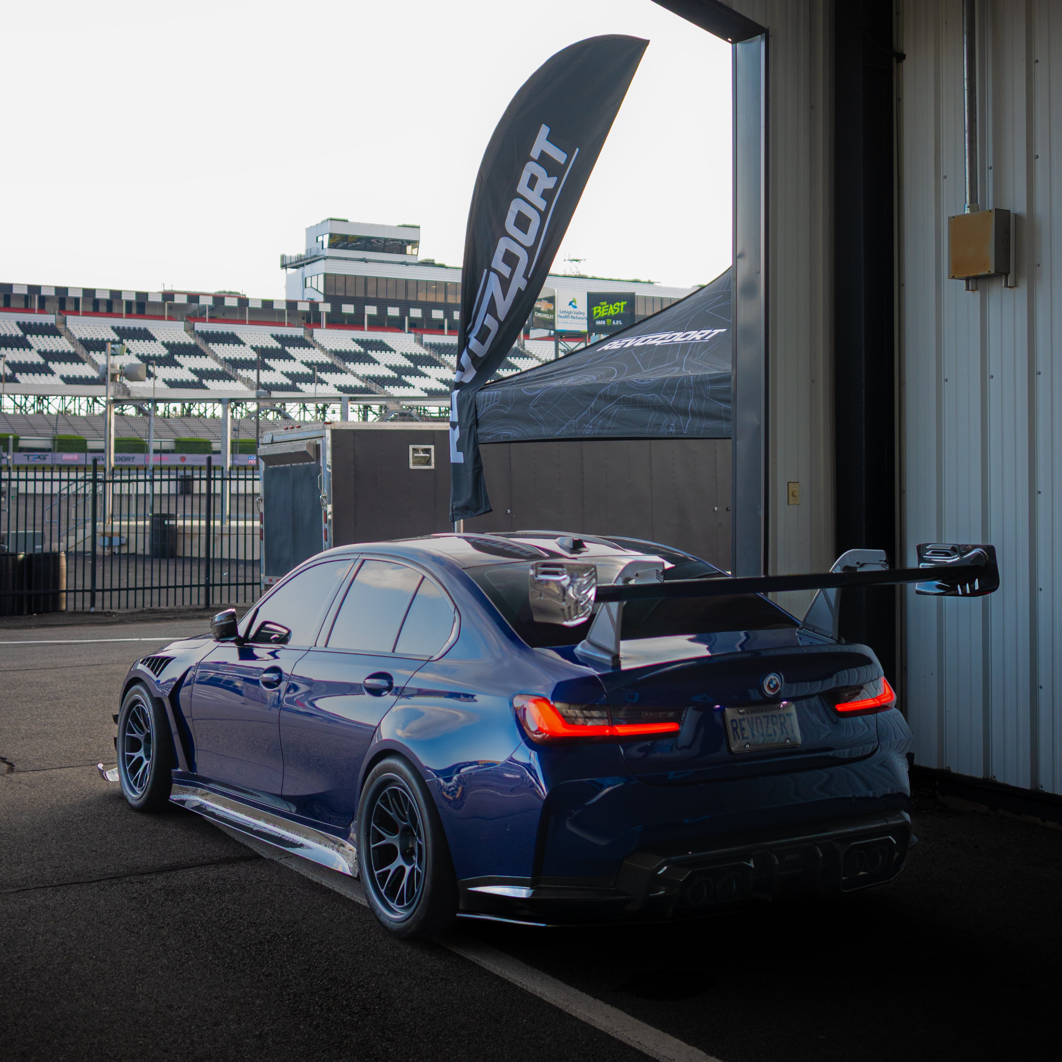 Blue BMW G80 M3 with aggressive carbon fiber aero kit, front splitter, vented fenders, and side skirts parked beside a professional race track during sunset.
