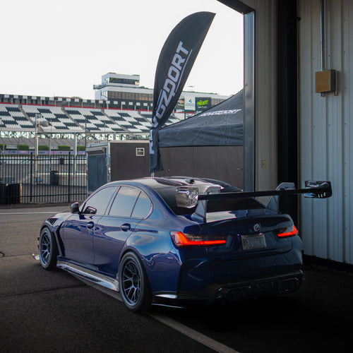Blue BMW G80 M3 with aggressive carbon fiber aero kit, front splitter, vented fenders, and side skirts parked beside a professional race track during sunset.