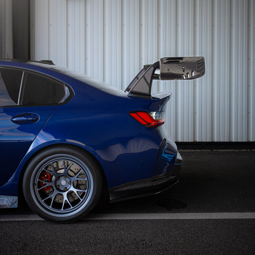 Blue BMW G80 M3 with aggressive carbon fiber aero kit, front splitter, vented fenders, and side skirts parked beside a professional race track during sunset.