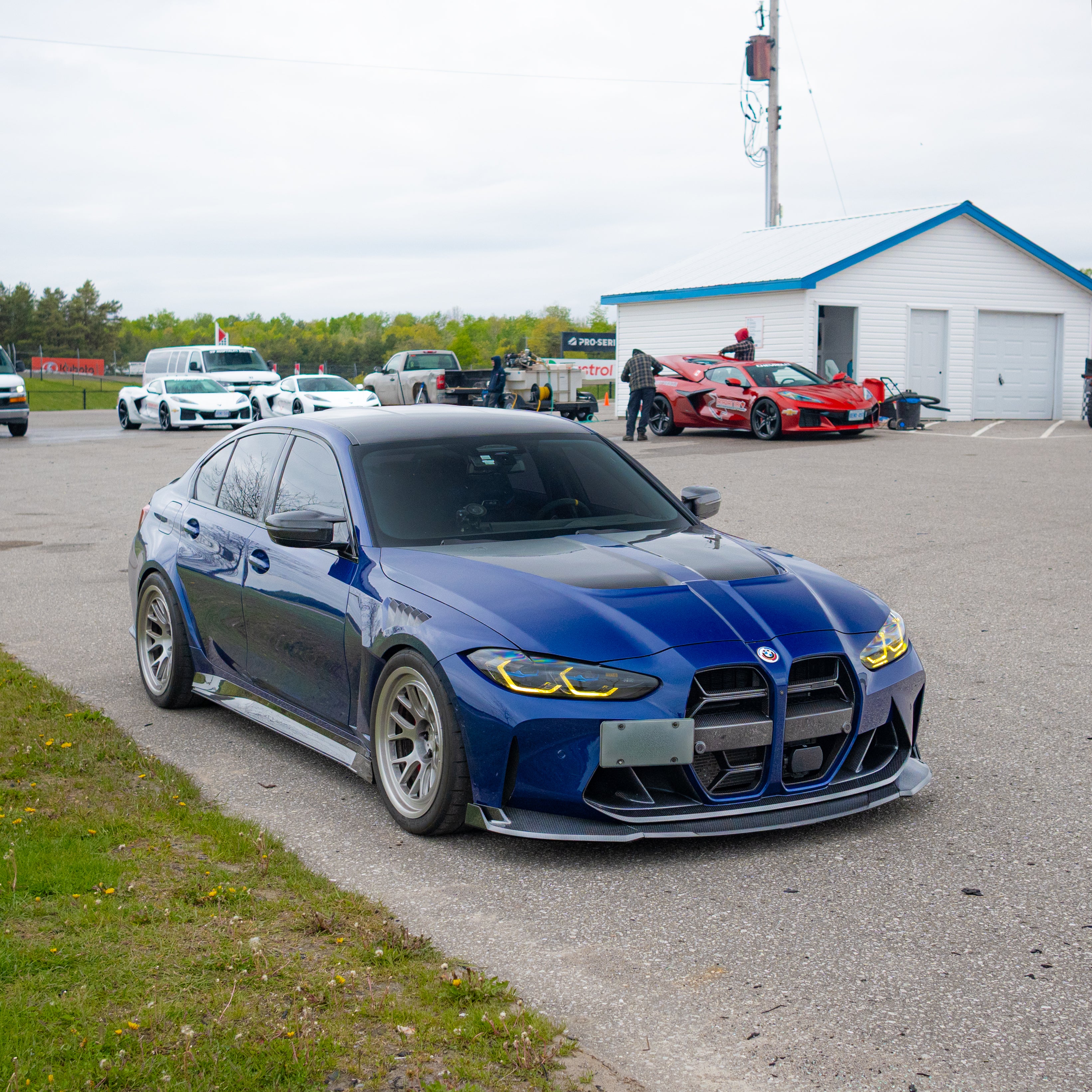 Blue BMW G80 M3 with aggressive carbon fiber aero kit, front splitter, vented fenders, and side skirts parked beside a professional race track during sunset.