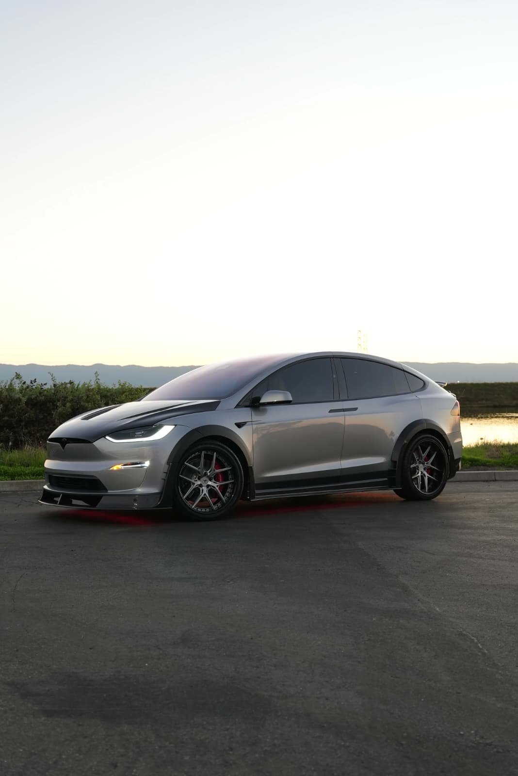 Tesla Model X featuring an aggressive REVOZPORT carbon fiber body kit with front lip, vented hood, wide fender extensions, side skirts, and illuminated red underglow lighting at dusk.