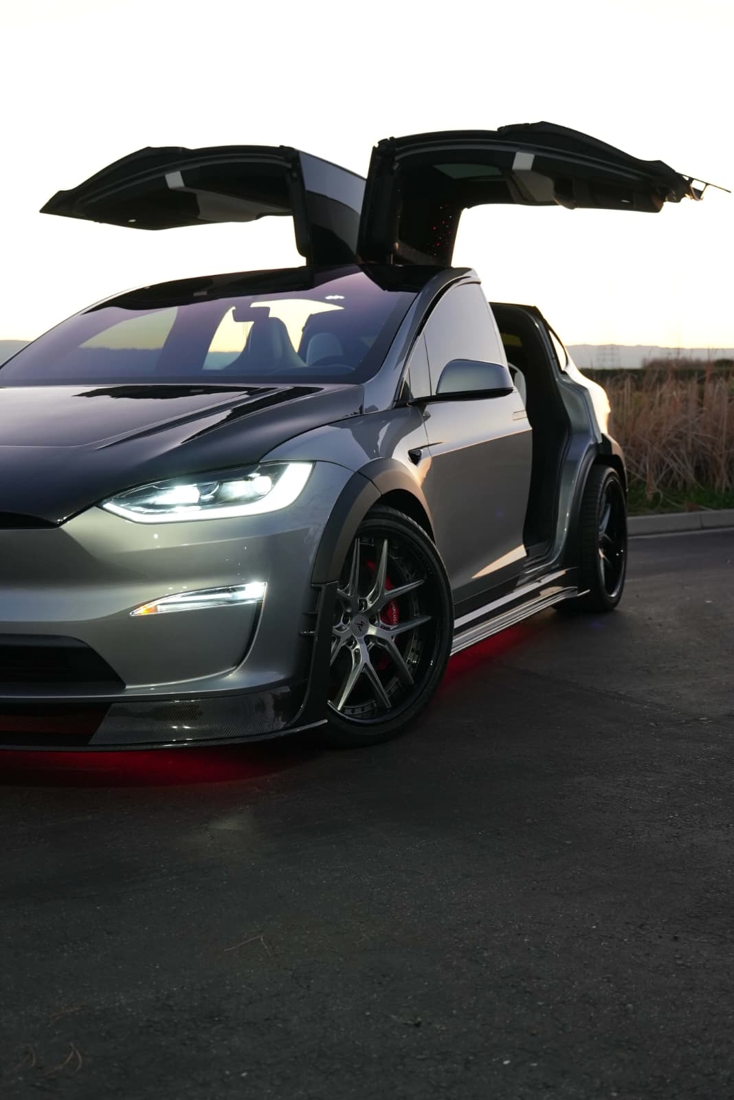 Tesla Model X featuring an aggressive REVOZPORT carbon fiber body kit with front lip, vented hood, wide fender extensions, side skirts, and illuminated red underglow lighting at dusk.