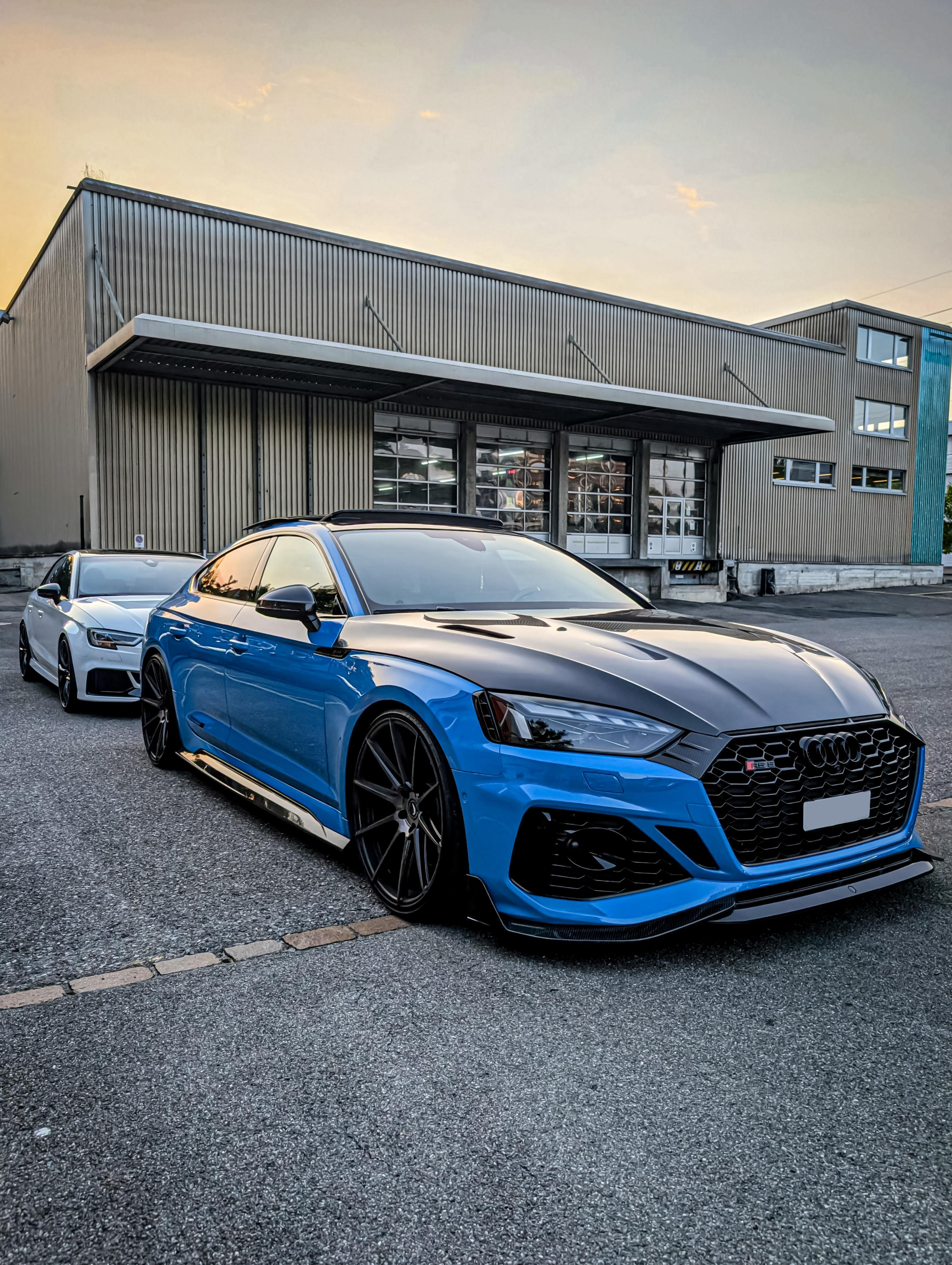 Blue Audi RS5 Sportback featuring a REVOZPORT carbon fiber body kit with vented hood, front lip, side skirts, and lowered stance parked outside a modern industrial building at sunset.