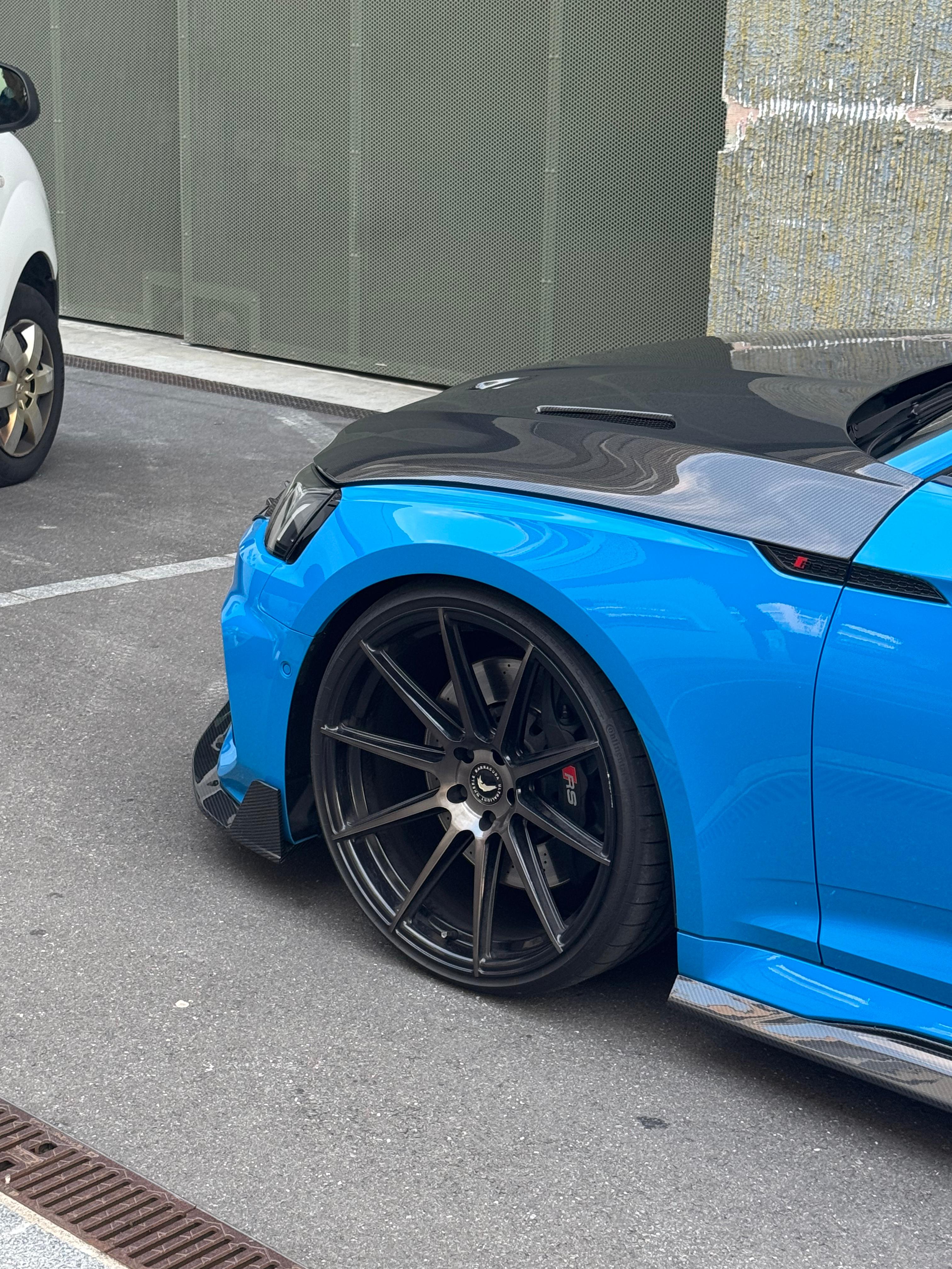 Blue Audi RS5 Sportback featuring a REVOZPORT carbon fiber body kit with vented hood, front lip, side skirts, and lowered stance parked outside a modern industrial building at sunset.