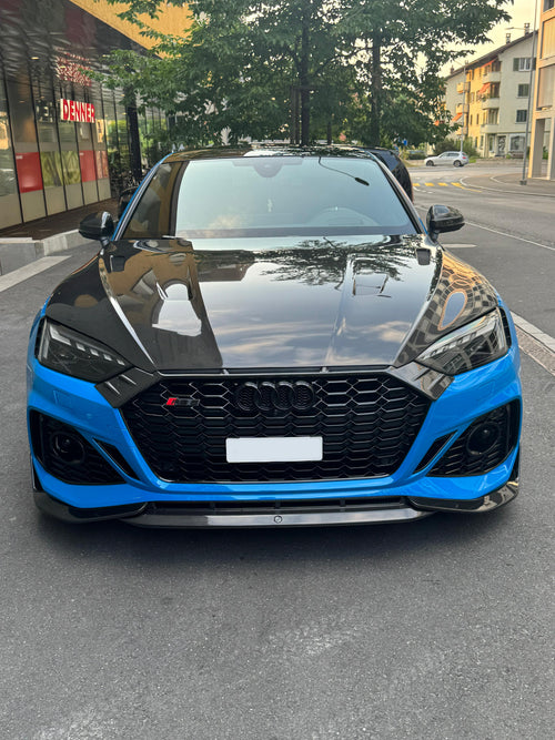 Blue Audi RS5 Sportback featuring a REVOZPORT carbon fiber body kit with vented hood, front lip, side skirts, and lowered stance parked outside a modern industrial building at sunset.
