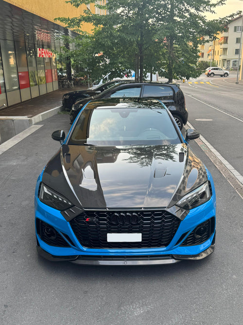 Blue Audi RS5 Sportback featuring a REVOZPORT carbon fiber body kit with vented hood, front lip, side skirts, and lowered stance parked outside a modern industrial building at sunset.