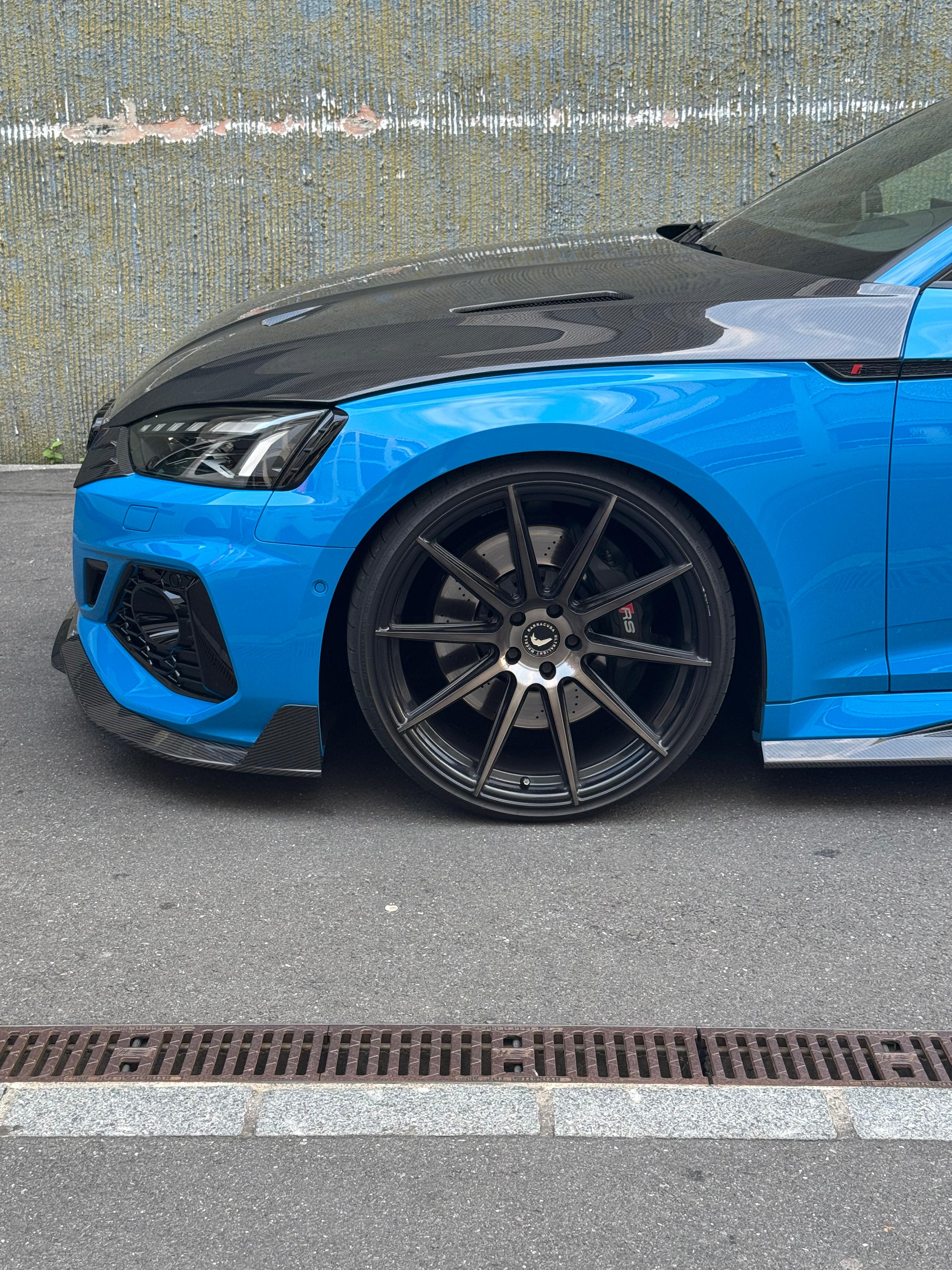 Blue Audi RS5 Sportback featuring a REVOZPORT carbon fiber body kit with vented hood, front lip, side skirts, and lowered stance parked outside a modern industrial building at sunset.