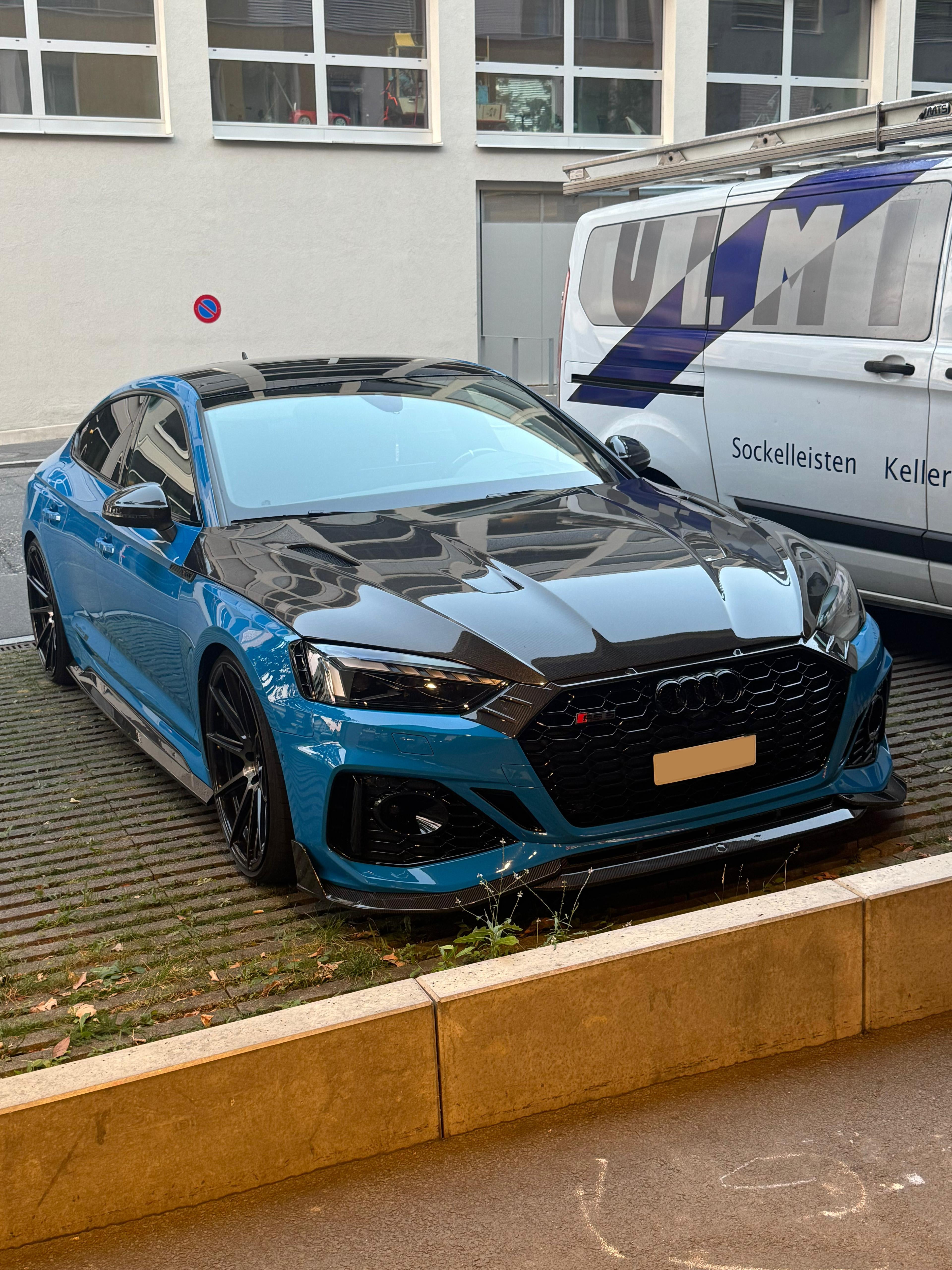 Blue Audi RS5 Sportback featuring a REVOZPORT carbon fiber body kit with vented hood, front lip, side skirts, and lowered stance parked outside a modern industrial building at sunset.