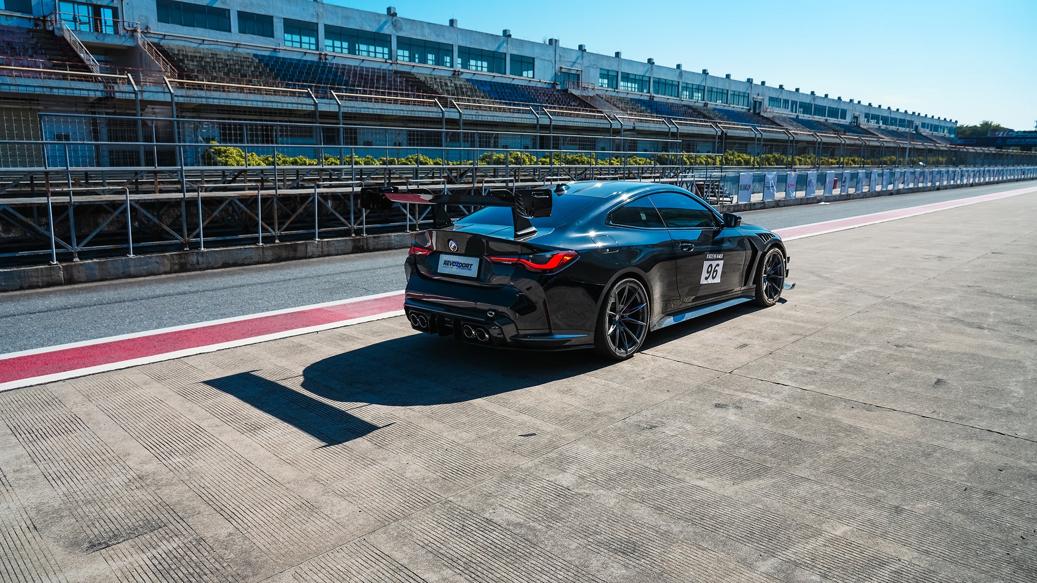 Black BMW G82 M4 track build with carbon fiber vented fenders, side skirts, rear diffuser, and oversized GT wing parked at a professional racing circuit pit lane.