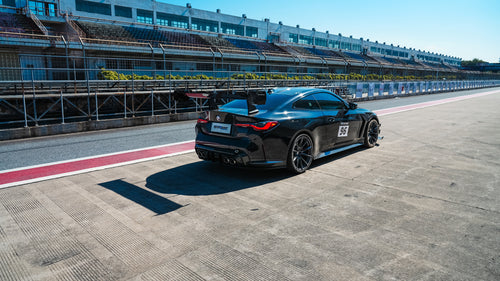 Black BMW G82 M4 track build with carbon fiber vented fenders, side skirts, rear diffuser, and oversized GT wing parked at a professional racing circuit pit lane.