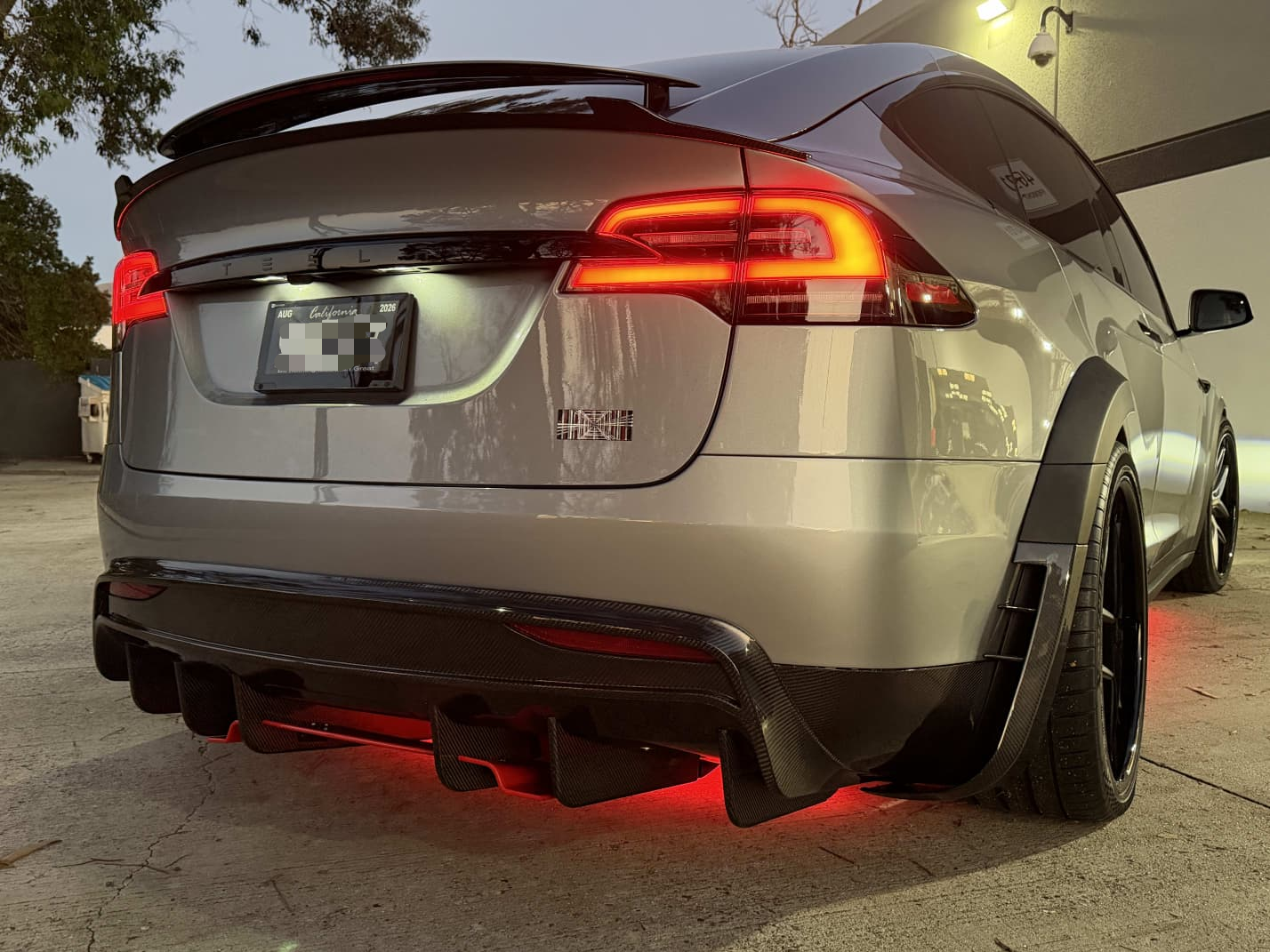 Tesla Model X featuring an aggressive REVOZPORT carbon fiber body kit with front lip, vented hood, wide fender extensions, side skirts, and illuminated red underglow lighting at dusk.