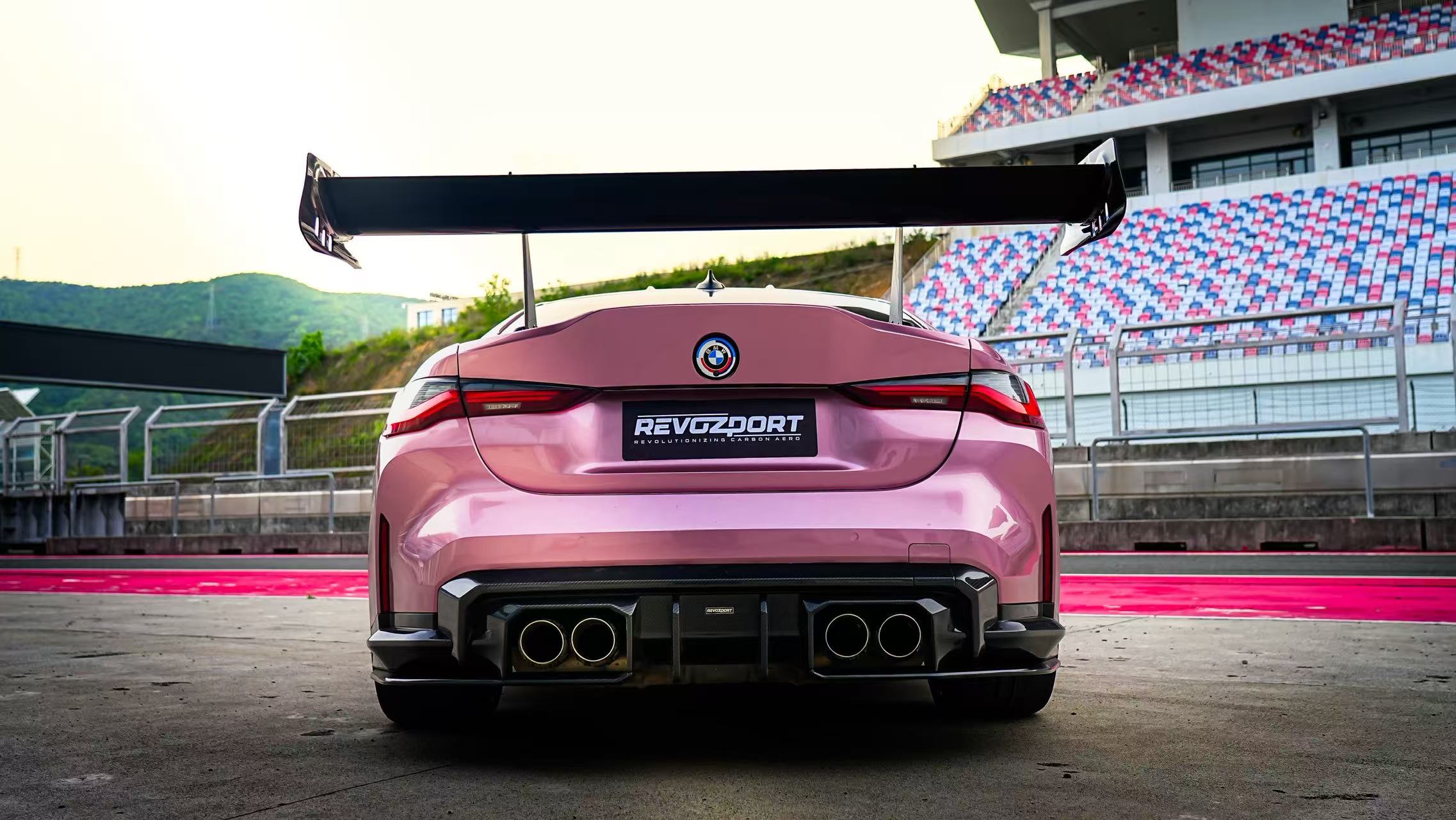 Pink BMW G82 M4 fitted with a REVOZPORT carbon fiber widebody aero kit, front splitter, hood vents, and side skirts on a race track pit lane.