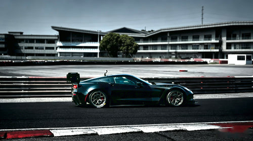 Black Chevrolet Corvette race car parked on a wet industrial track surface, featuring an aggressive wide-body kit, large rear wing, carbon fiber aerodynamic components, and lightweight performance wheels with red brake calipers. The moody urban backdrop and reflective pavement emphasize the car’s high-performance motorsport styling.