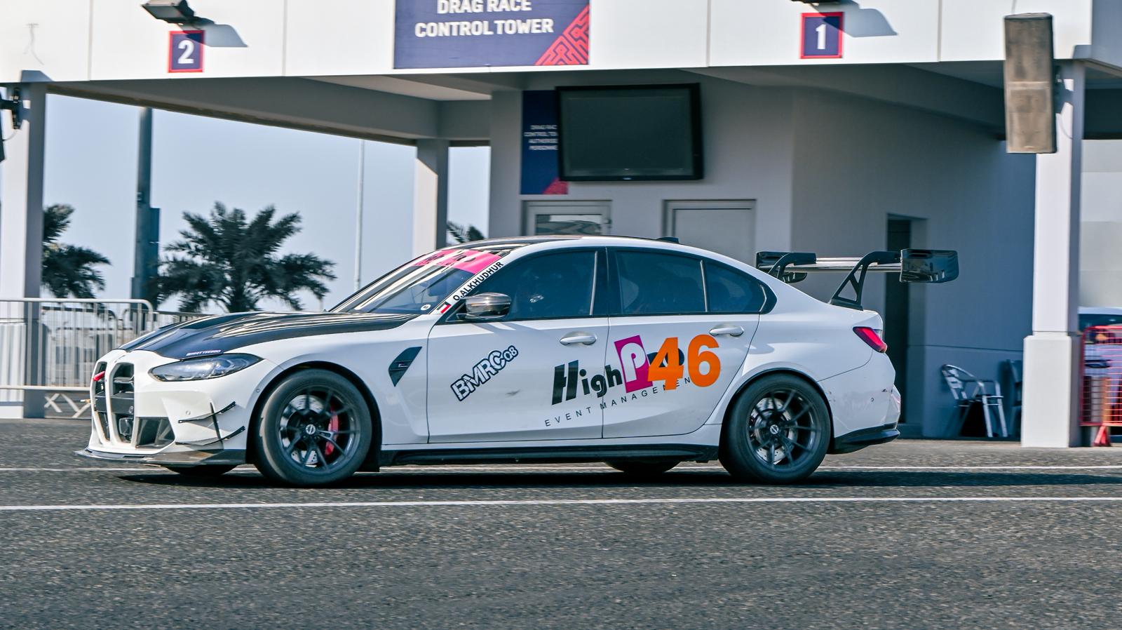 White BMW G80 M3 track car with REVOZPORT carbon fiber aero kit, front canards, rear wing, and racing decals driving on a professional race circuit.