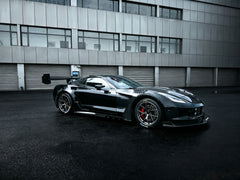 Black Chevrolet Corvette race car parked on a wet industrial track surface, featuring an aggressive wide-body kit, large rear wing, carbon fiber aerodynamic components, and lightweight performance wheels with red brake calipers. The moody urban backdrop and reflective pavement emphasize the car’s high-performance motorsport styling.