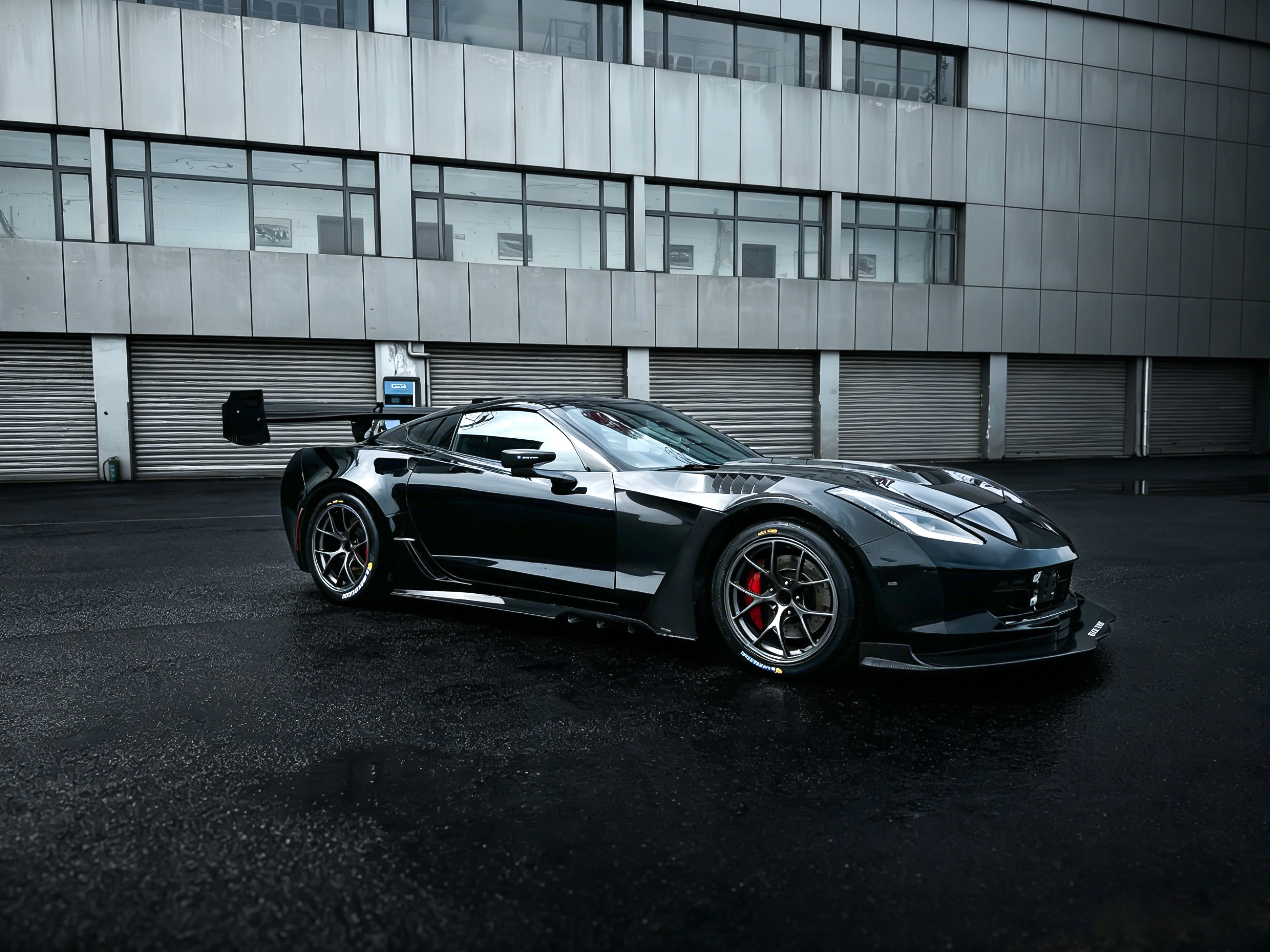 Black Chevrolet Corvette race car parked on a wet industrial track surface, featuring an aggressive wide-body kit, large rear wing, carbon fiber aerodynamic components, and lightweight performance wheels with red brake calipers. The moody urban backdrop and reflective pavement emphasize the car’s high-performance motorsport styling.