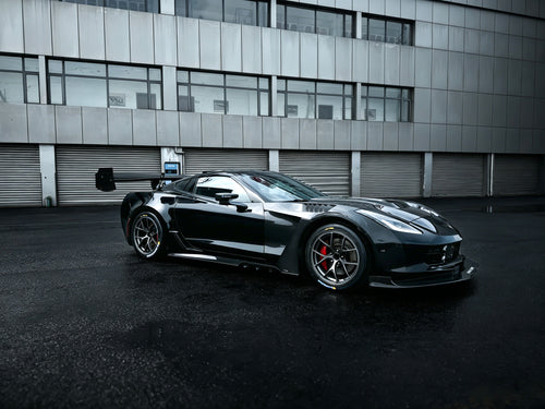 Black Chevrolet Corvette race car parked on a wet industrial track surface, featuring an aggressive wide-body kit, large rear wing, carbon fiber aerodynamic components, and lightweight performance wheels with red brake calipers. The moody urban backdrop and reflective pavement emphasize the car’s high-performance motorsport styling.