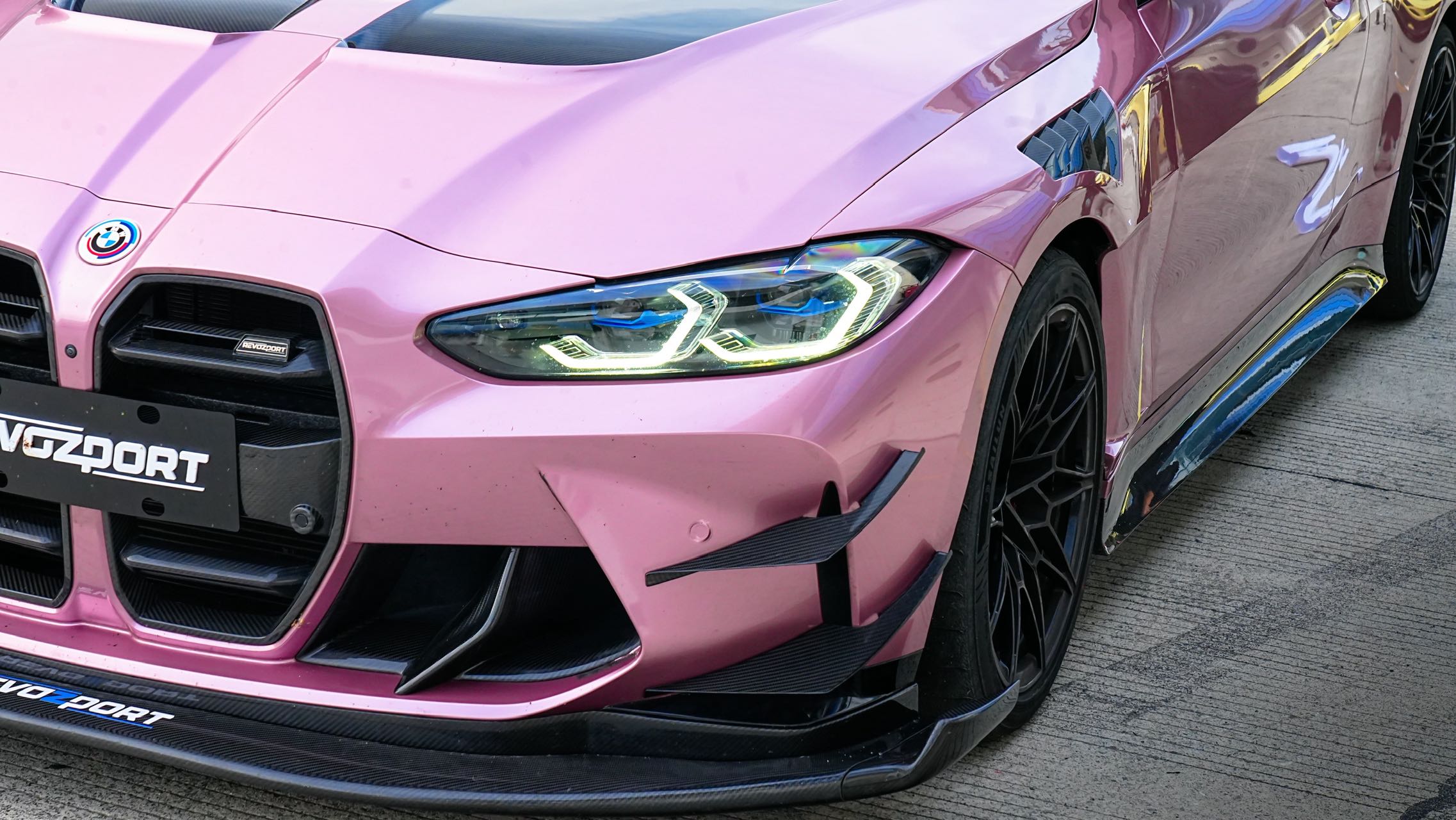 Pink BMW G82 M4 fitted with a REVOZPORT carbon fiber widebody aero kit, front splitter, hood vents, and side skirts on a race track pit lane.