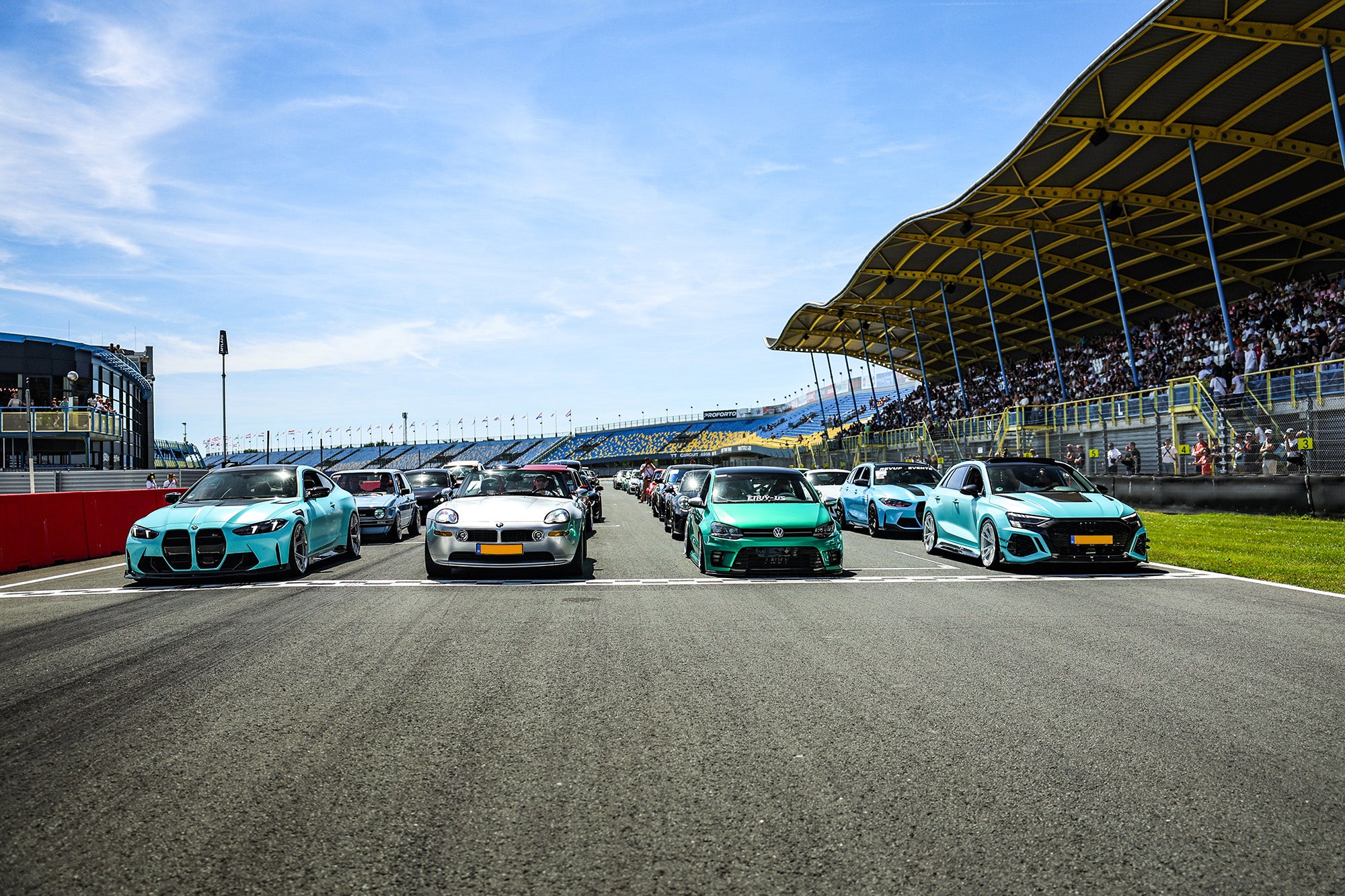 Multiple modified performance cars lined up on a racetrack grid, showcasing carbon fiber aero styling at a motorsport event.