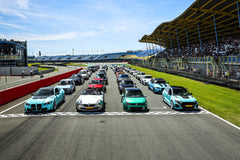 Multiple modified performance cars lined up on a racetrack grid, showcasing carbon fiber aero styling at a motorsport event.