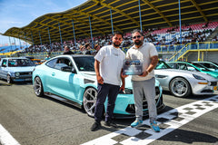 Turquoise BMW with carbon fiber aero kit displayed on a racetrack, with two owners holding a show award.