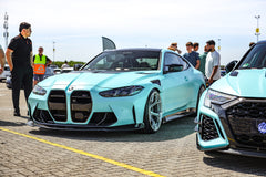 Turquoise BMW with carbon fiber aero kit displayed at a sunny car event, with visitors interacting beside the car.