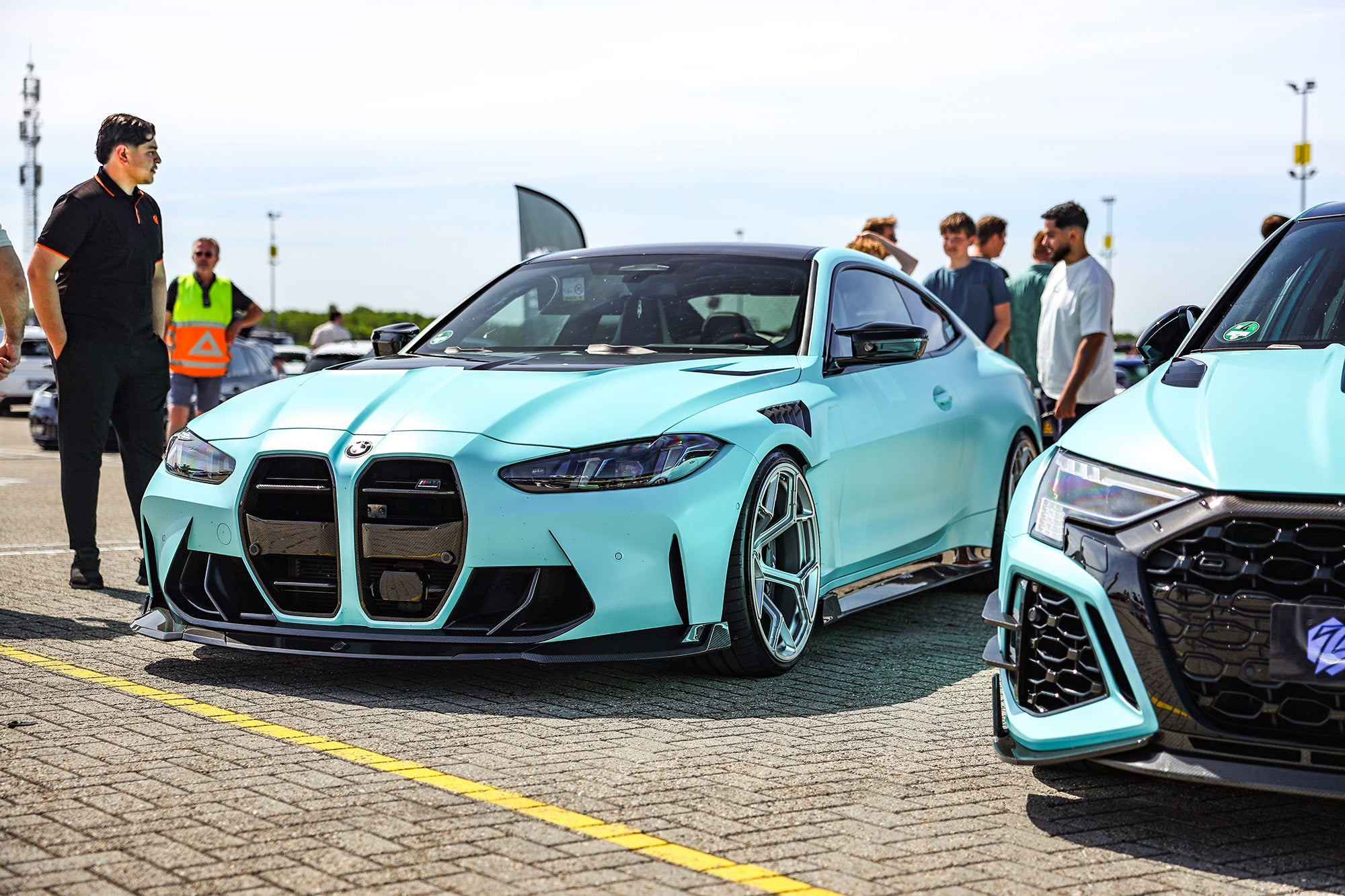 Turquoise BMW with carbon fiber aero kit displayed at a sunny car event, with visitors interacting beside the car.