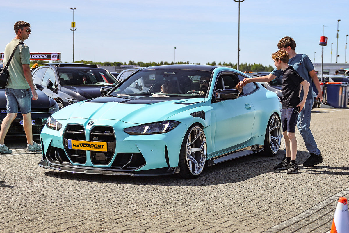 Turquoise BMW with carbon fiber aero kit displayed at a sunny car event, with visitors interacting beside the car.