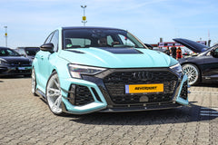 Turquoise Audi with carbon fiber body kit and white wheels displayed at an outdoor car event, highlighting aggressive front aero styling.