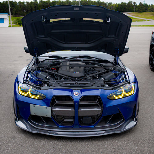 Blue BMW G80 M3 with aggressive carbon fiber aero kit, front splitter, vented fenders, and side skirts parked beside a professional race track during sunset.