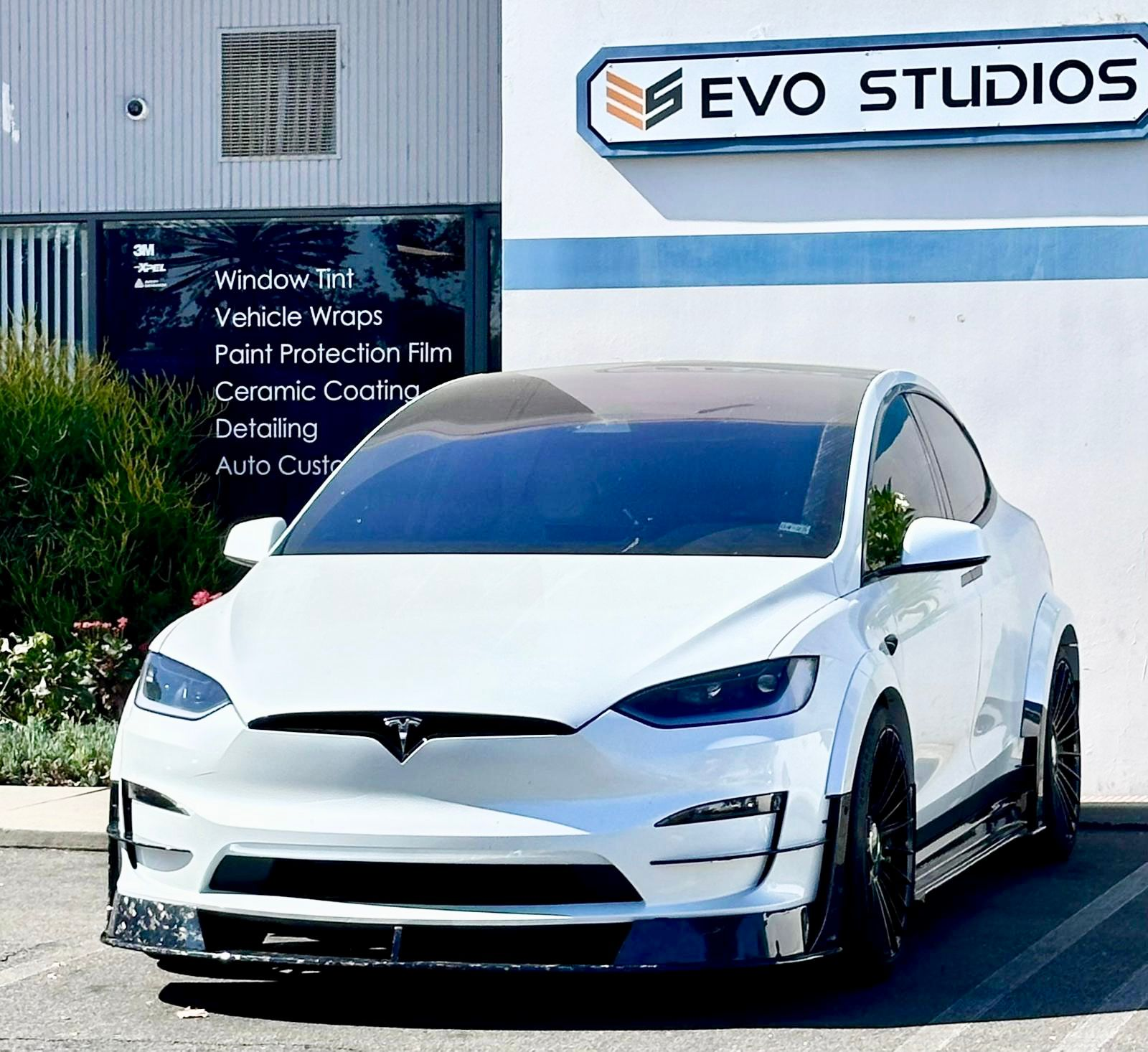 White Tesla Model X featuring an aggressive REVOZPORT body kit with carbon fiber front lip, side skirts, wide fender extensions, and lowered stance parked outside a detailing studio.